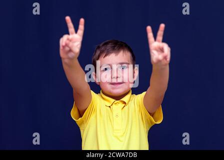 ragazzo con le dita mostra la pace o la vittoria segno su blu sfondo scuro dello studio Foto Stock