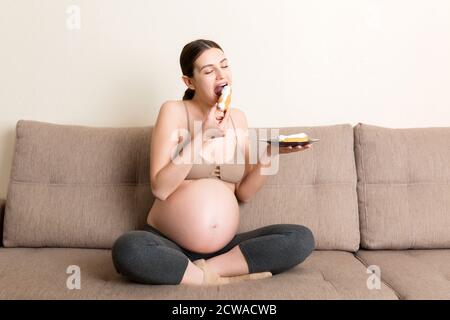 La donna incinta affamata sta mangiando la pasticceria con grande piacere rilassarsi sul divano a casa. Buon concetto di gravidanza. Foto Stock