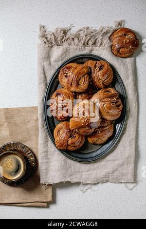Cannella tradizionale svedese confetti dolci Kanelbulle su vassoio vintage, tazza di caffè, bastoncini di cannella su tessuto di lino su sfondo bianco texture. Piatto Foto Stock
