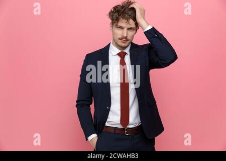 giovane uomo d'affari indecorato in tuta blu navy tenendo la mano in tasca e graffiando la testa, pensando e in piedi su sfondo rosa Foto Stock