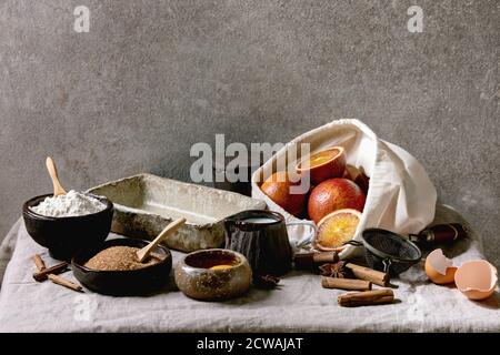 Ingredienti per la cottura a casa. Farina, uova, zucchero di canna, latte in diverse ciotole di ceramica, arance di sangue e spezie su tela da tavola di lino. Struttura grigia bac Foto Stock