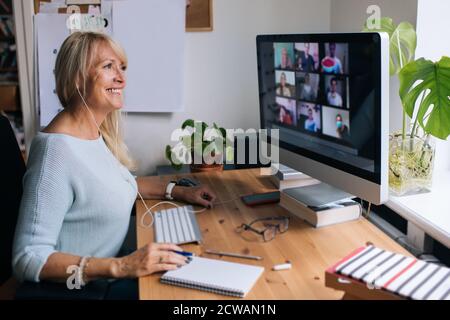 Donna matura sorridente che ha videochiamata via computer l'ufficio domestico. Videoconferenza online per riunioni di team da casa. Donna d'affari attraente Foto Stock