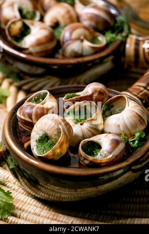Escargots de Bourgogne - lumache con burro alle erbe, piatto gourmet, in due tradizionali padelle in ceramica con coriandolo e pane su tovagliolo di paglia. Primo piano Foto Stock