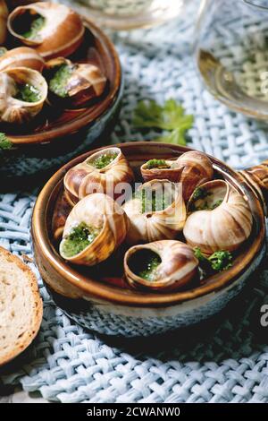 Escargots de Bourgogne - lumache con burro alle erbe, piatto gourmet, in tradizionale padella di ceramica con coriandolo, pane, bicchiere di vino bianco su paglia blu n Foto Stock