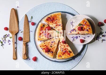 Cheesecake fatta in casa con lamponi al forno a fette su piatto decorato da lamponi freschi, fiori commestibili e menta con server di torta in legno e coltello rotondo Foto Stock