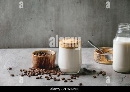 Dalgona Frothy caffè tendenza bevanda coreana latte latte latte con schiuma di caffè in tazza di vetro, decorato da caffè macinato su tavola grigia texture. Ingredienti sopra Foto Stock