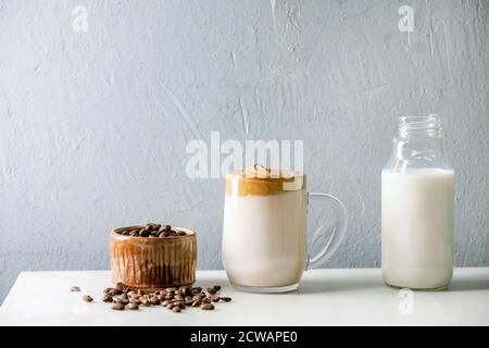 Dalgona Frothy caffè tendenza bevanda coreana latte latte latte con schiuma di caffè in tazza di vetro con ingredienti in fila su tavolo di marmo bianco. Foto Stock