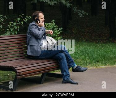 Mosca/Russia - Set 2020: Uomo in abiti casual con portatile e telefono cellulare seduto in panchina al parco della città per una conversazione Foto Stock