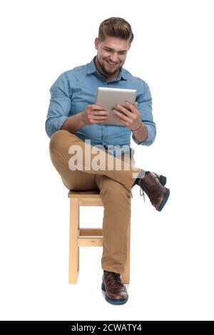 uomo giovane hapy in camicia di denim sorridente e tenendo linguetta, seduta isolata su sfondo bianco, corpo pieno Foto Stock
