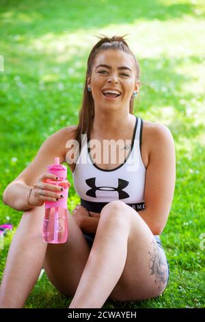 Una giovane donna che indossa abiti sportivi seduti all'esterno sull'erba tenendo una bottiglia d'acqua rosa e ridendo. Foto Stock