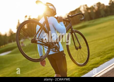 Forte atletico uomo in abbigliamento sportivo e casco protettivo che porta la sua bicicletta dopo l'allenamento in bicicletta nel parco, godendo di un incredibile tramonto. Stile di vita attivo e concetto sportivo. Escursioni in bicicletta all'aperto Foto Stock