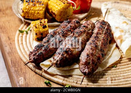 Fast food orientale. Lyulya kebab di manzo piccante alla griglia su bastoncini di pane piatto con verdure grigliate, pannocchie di mais dolci, pomodoro e paprika, salsa di pomodoro Foto Stock