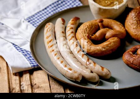 Tradizionale salsiccia di maiale alla griglia bavarese su piatto di ceramica servita con senape dolce tedesca e pretzel pane su bianco e blu tovagliolo su legno b Foto Stock