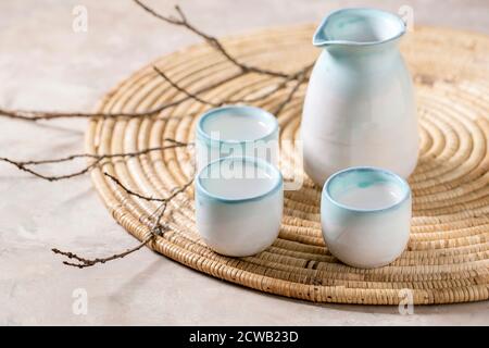 Set in ceramica sake per il tradizionale vino di riso con bevande alcoliche giapponesi, boccetta e tre tazze, in piedi su tovagliolo di paglia con rami asciutti su beige Foto Stock