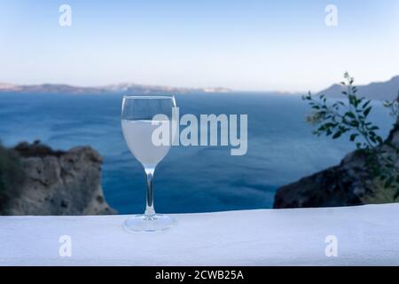 Primo piano di un vetro con gambo ouzo su un bianco Ammira il tramonto con la caldera di Santorini e il mare blu sullo sfondo Foto Stock