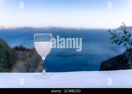 Primo piano di un vetro con gambo ouzo su un bianco Ammira il tramonto con la caldera di Santorini e il mare blu sullo sfondo Foto Stock