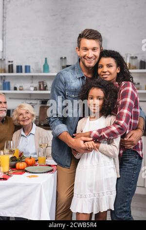 Fuoco selettivo dei genitori che abbracciano la figlia afroamericana vicino al cibo sul tavolo durante il ringraziamento Foto Stock
