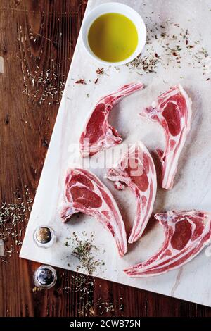 Materie costolette di agnello con sale e pepe, asciugare le erbe aromatiche e la coppa dell' olio d' oliva sul marmo scheda sopra un tavolo di legno. Vista dall'alto. Foto Stock