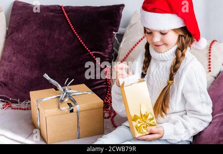 Una ragazza in un cappello di babbo natale si siede sul divano e apre un regalo di nuovo anno in un oro pacchetto Foto Stock