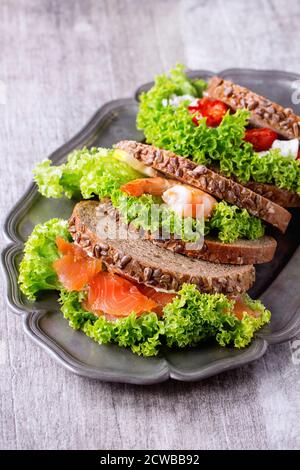 Set di panini con pane integrale, insalata fresca, formaggio feta, pomodori ciliegini, gamberi e salmone salato su piatto di metallo vintage su legno bianco Foto Stock