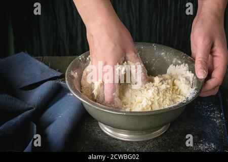 Rendendo la pasta frolla impasto da donna con le mani in mano in vintage recipiente metallico sopra il vecchio tavolo in legno. Scuro in stile rustico Foto Stock