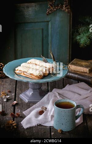 Vecchio tavolo di Natale in legno con crema di burro Eclair e tazza di tè caldo, decorando da stelle di Natale, albero e libri d'annata. Con la vecchia porta aperta in Foto Stock