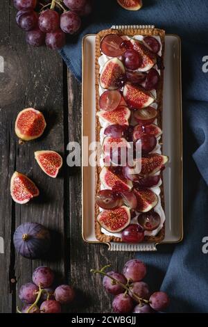 Crostata rettangolare fatta in casa con uva rossa, fichi e panna montata in piastra di ceramica bianca su vecchio tavolo di legno con straccio di tessuto blu. Stile rustico scuro Foto Stock