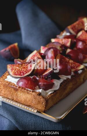 Crostata rettangolare fatta in casa con uva rossa, fichi e panna montata in piastra di ceramica bianca su vecchio tavolo di legno con straccio di tessuto blu. Stile rustico scuro Foto Stock