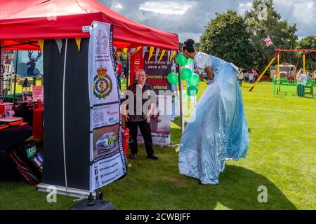 Una ragazza intrattenitrice su palafitte parla ad UN titolare di stall al Hartfield Village Fete, Hartfield, East Sussex, Regno Unito. Foto Stock