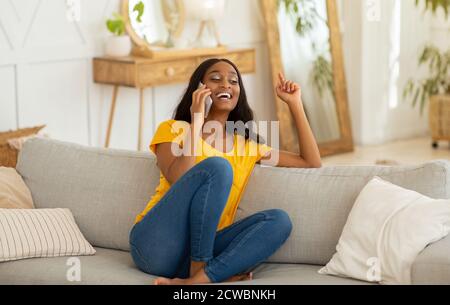 Donna afro-americana di charme che parla su smartphone e ridendo divano a casa Foto Stock