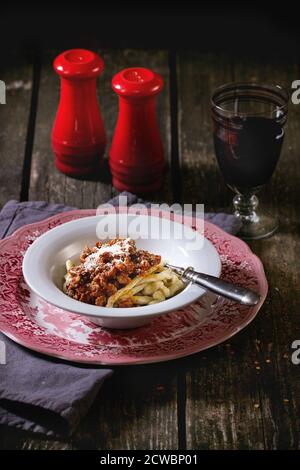 Piatto inglese d'epoca di pasta pici fatta in casa con salsa bolognese, servito su tovagliolo grigio con bicchiere di vino rosso, sale rosso e pepi shakers o Foto Stock