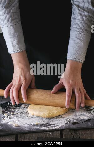 Mani femminili rolling out impasto per la pasta di legno mattarello in legno tavolo da cucina, polverizzazione da farina. Scuro in stile rustico. Foto Stock