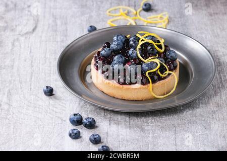 Round crostata al limone con frutta fresca e cotta mirtilli, servita su vintage piastra metallica con la scorza di limone su grigio Sfondo di legno. Foto Stock