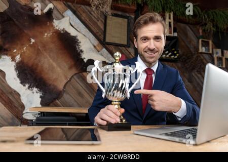 uomo d'affari orgoglioso che punta il dito al trofeo e sorridente, seduto in un coffee shop Foto Stock