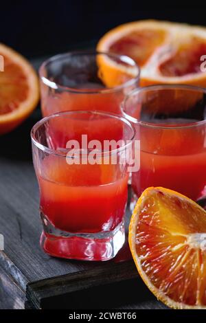 Primo piano di Shorts di cocktail alcolici con arance di sangue siciliano a fette e succo d'arancia rosso fresco, serviti su tavolo di legno nero. Stile rustico scuro. Foto Stock