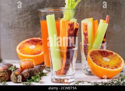 Affettato carote e sedano crudi colorate come spuntino vegetariano, arance e timo di sangue con una tazza di bicchiere di succo di arancia e carota fresco su pietra bianca t Foto Stock