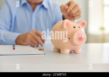 Uomo anziano risparmiando soldi e mettendo moneta in cute lucida banco di porcellini in piedi sulla scrivania Foto Stock