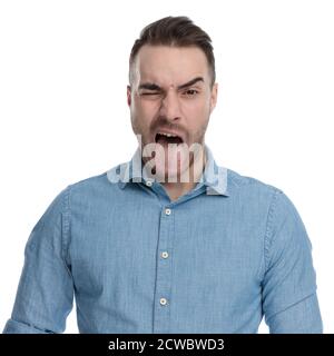 Divertente uomo casual che si strinca e si stacca la lingua mentre indossa una camicia blu, in piedi su sfondo bianco studio Foto Stock