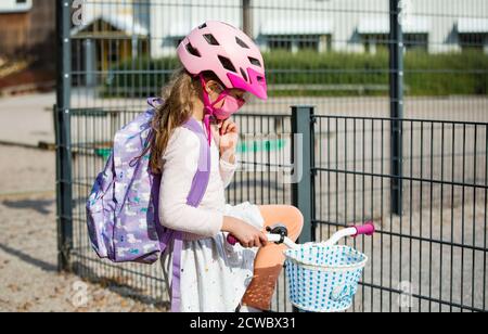 Studentessa indossa un tessuto protettivo maschera riutilizzabile che cavalca una bicicletta a scuola. Educazione scolastica durante la pandemia. Distanza sociale Foto Stock