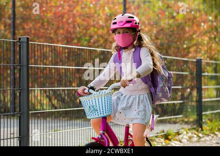 Studentessa indossa un tessuto protettivo maschera riutilizzabile che cavalca una bicicletta a scuola. Educazione scolastica durante la pandemia. Distanza sociale Foto Stock