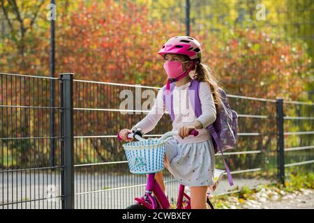 Studentessa indossa un tessuto protettivo maschera riutilizzabile che cavalca una bicicletta a scuola. Educazione scolastica durante la pandemia. Distanza sociale Foto Stock