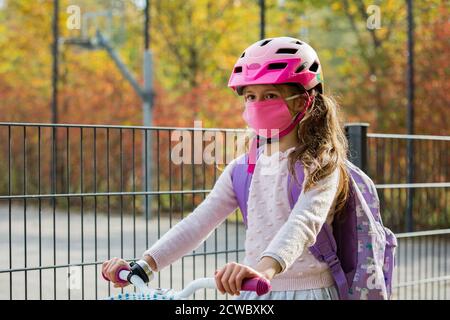 Studentessa indossa un tessuto protettivo maschera riutilizzabile che cavalca una bicicletta a scuola. Educazione scolastica durante la pandemia. Distanza sociale Foto Stock