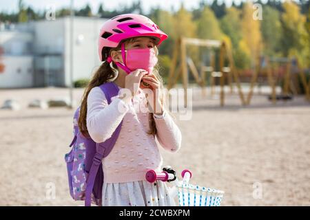 Studentessa indossa un tessuto protettivo maschera riutilizzabile che cavalca una bicicletta a scuola. Educazione scolastica durante la pandemia. Distanza sociale Foto Stock