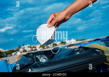 closeup di un uomo caucasico che lancia un usato bianco monouso maschera al cestino vicino alla spiaggia Foto Stock