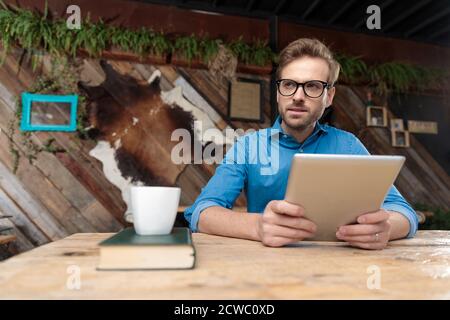 uomo casual che indossa occhiali seduti alla scrivania e che lo tiene in mano tablet mentre si guarda via pusivo al caffè Foto Stock