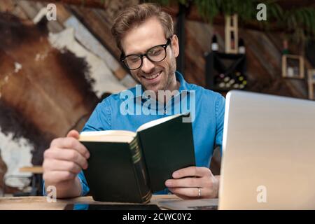 uomo informale che indossa occhiali seduti alla scrivania che studia dal libro felice al coffeeshop Foto Stock