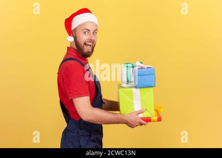 Vista laterale uomo di consegna estremamente felice in uniforme blu e cappello rosso di babbo natale che tiene le scatole presenti guardando la macchina fotografica con gli occhi grandi e la bocca aperta. IO Foto Stock