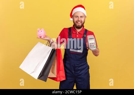 Consegna positiva uomo in uniforme e cappello di babbo natale che tiene borse di carta shopping, terminale di pagamento e banca piggy, pagando contactless e risparmiando soldi Foto Stock