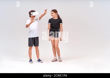 Tecnologia VR. Foto a tutta lunghezza di un ragazzo adolescente che indossa occhiali per realtà virtuale e una ragazza carina in piedi isolata su sfondo grigio Foto Stock