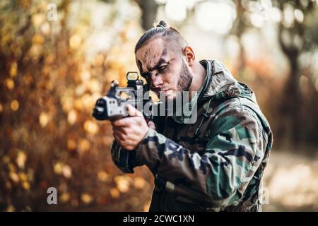 Un soldato in uniforme camouflage e volto dipinto è mirato. Tiene un fucile. Foto a mezza altezza. Airsoft. Foto Stock